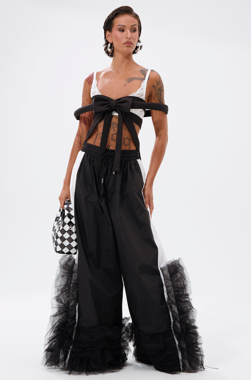 A person with short hair and tattoos models the IM THE GIFT CHEST PIECE sheer top, paired with wide black pants featuring tulle ruffle hems, holding a black-and-white checkered bag against a plain white backdrop. 3