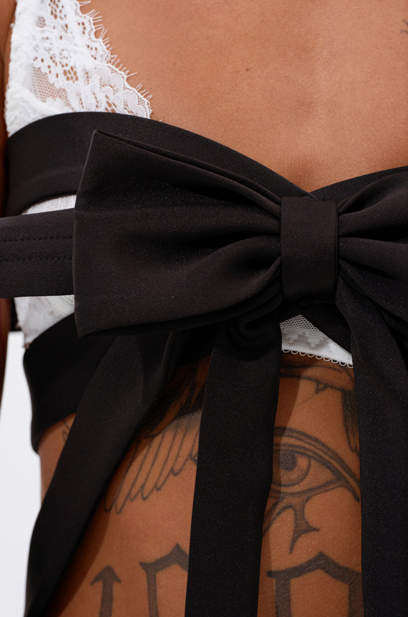 Close-up of a person wearing the IM THE GIFT CHEST PIECE, an exaggerated bow chest accessory styled over a white lace bralette, with tattoos visible beneath the bold silhouette. 4