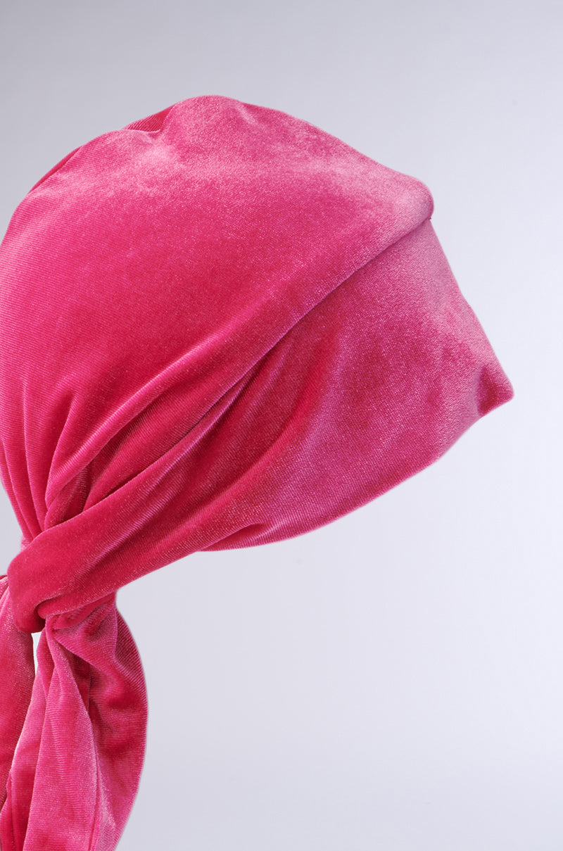 A close-up side view of someone wearing a bright pink RAY VELVET DURAG, tied at the back, with most of their face covered by the durag and set against a plain light gray background.
