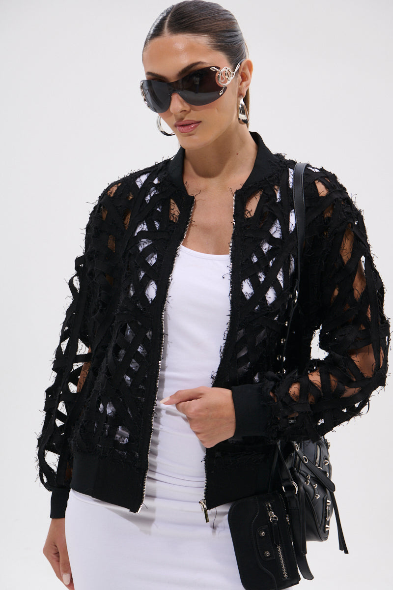 A woman poses confidently against a plain background wearing the IONA CUTOUT DENIM BOMBER, oversized sunglasses, hoop earrings, a white dress, and holding a black handbag.