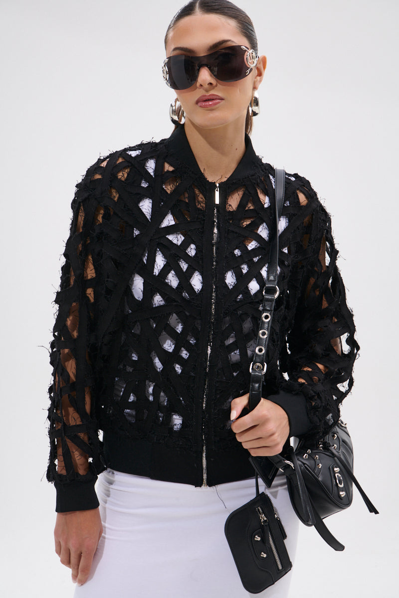 A woman wearing large sunglasses, hoop earrings, and the IONA CUTOUT DENIM BOMBER over a white top and skirt holds a black purse with a silver chain, standing against a plain white background.