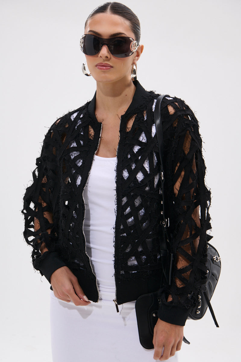 A woman wearing large black sunglasses, hoop earrings, a white top and skirt, and the IONA CUTOUT DENIM BOMBER with a lattice-patterned sheer design stands against a plain white background holding a black bag.
