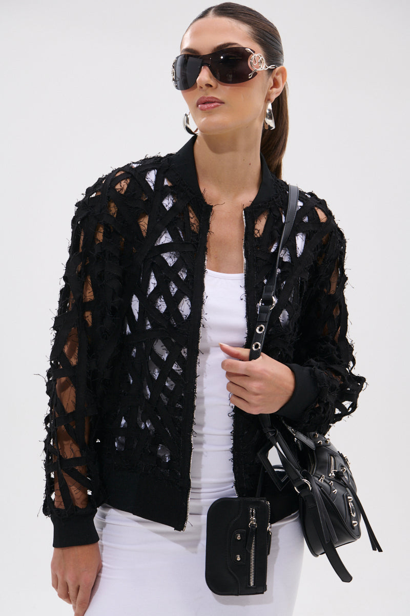 A woman in the IONA CUTOUT DENIM BOMBER over a white top and pants, with oversized sunglasses and two black crossbody bags, stands against a plain light background.