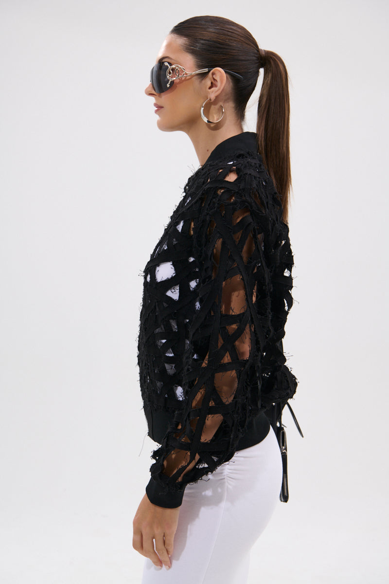 Woman in profile wears the IONA CUTOUT DENIM BOMBER—an oversized black denim sheer jacket with crisscrossed raw edges and cut-out details—paired with large sunglasses, hoop earrings, and white pants against a plain white background.