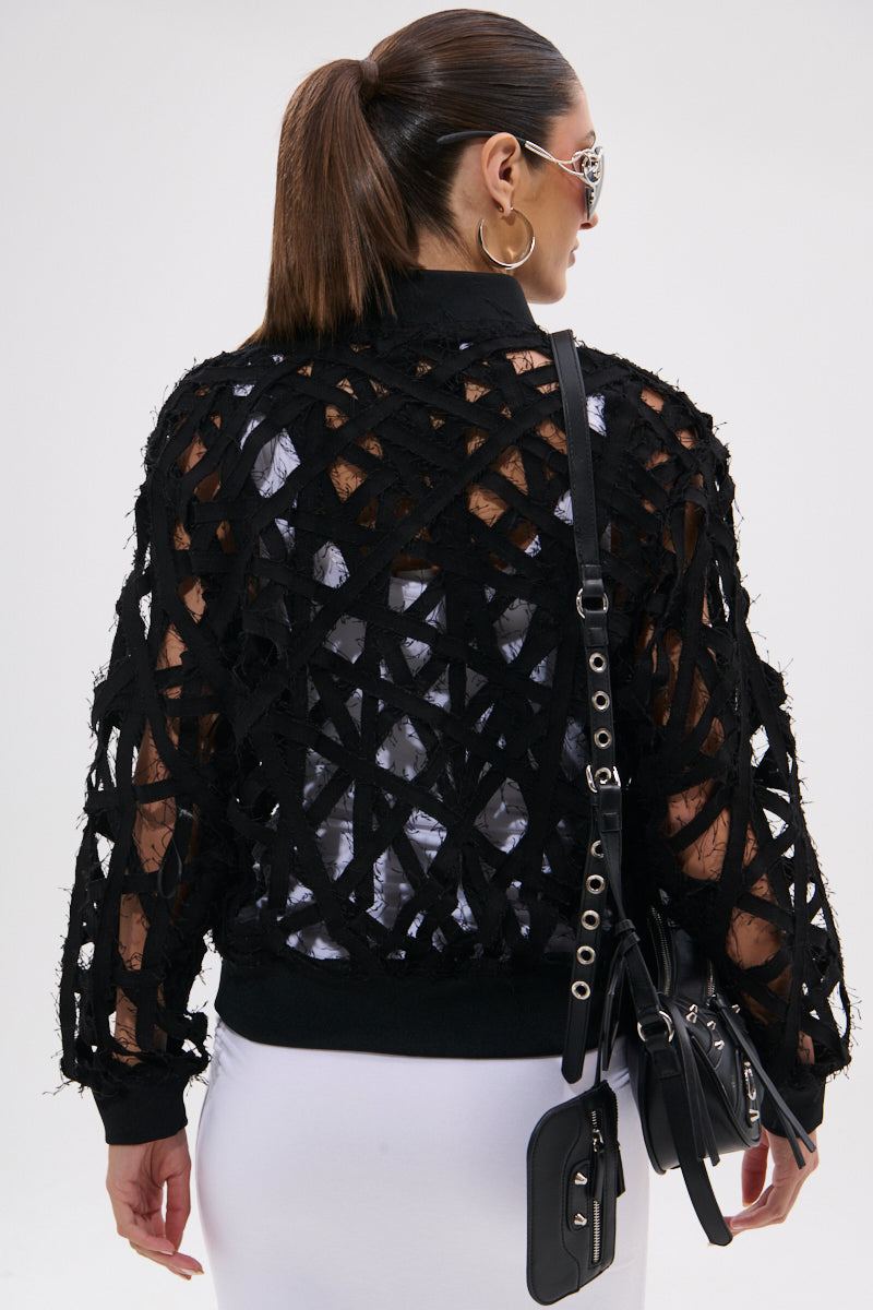 A woman with a ponytail, sunglasses, and hoop earrings is seen from the back wearing the IONA CUTOUT DENIM BOMBER over a white top and skirt. She carries a black crossbody bag with silver hardware.