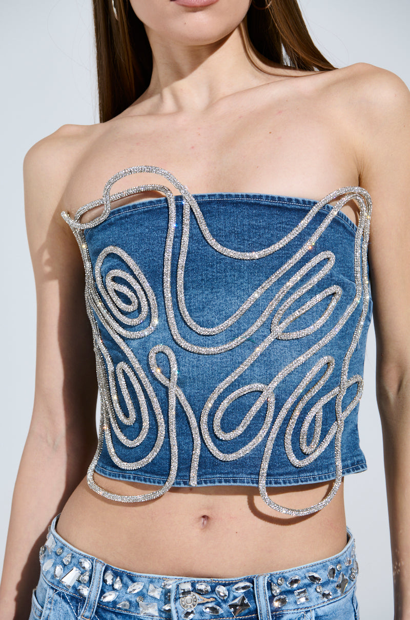 A person showcases the WOBBLY RHINESTONE DENIM CORSET TOP, featuring abstract silver embellishments. It's paired with rhinestone denim jeans against a plain white backdrop, with their long hair partially visible, highlighting the outfit's edgy elegance.