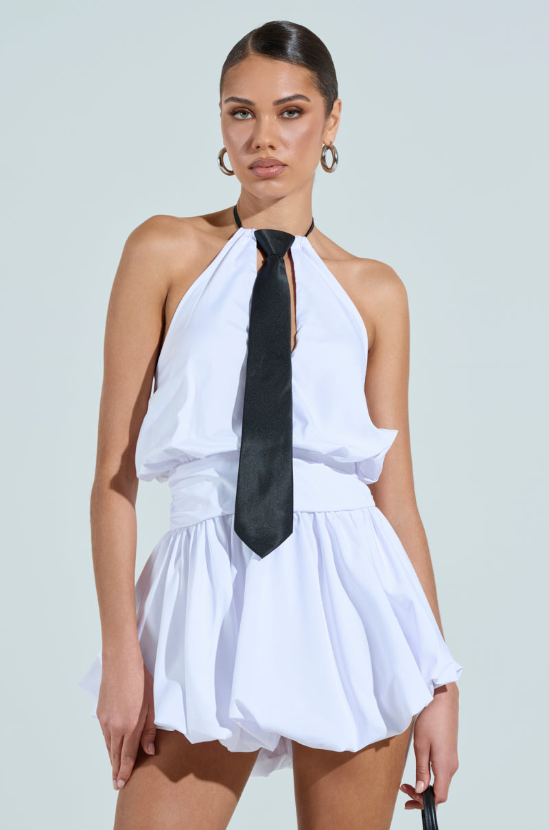 A woman wears the OUT OF OFFICE MINI BUBBLE DRESS WITH TIE, a sleeveless white romper with a halter neckline, loose skirt, and black tie. Her hair is slicked back; she accessorizes with large hoop earrings and poses against a plain light background.