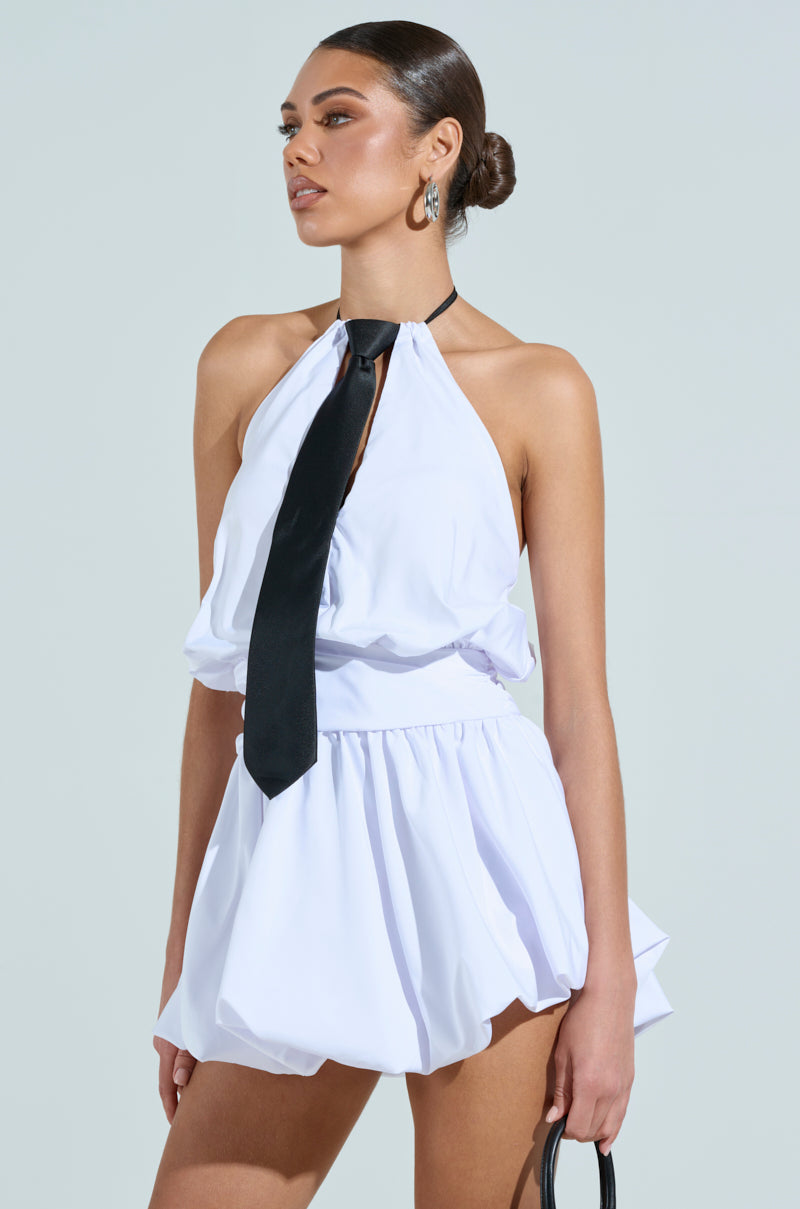 A woman stands confidently in the OUT OF OFFICE MINI BUBBLE DRESS WITH TIE, featuring a white halter neckline, cinched waist, black necktie, and styled with a sleek bun and small black handbag.