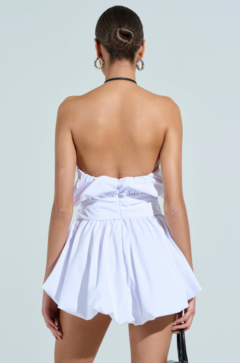 A woman with a sleek bun wears the OUT OF OFFICE MINI BUBBLE DRESS WITH TIE, a white, strapless, backless mini with a cinched waist and flared skirt. She carries a small black bag and stands facing away against a plain light gray background.