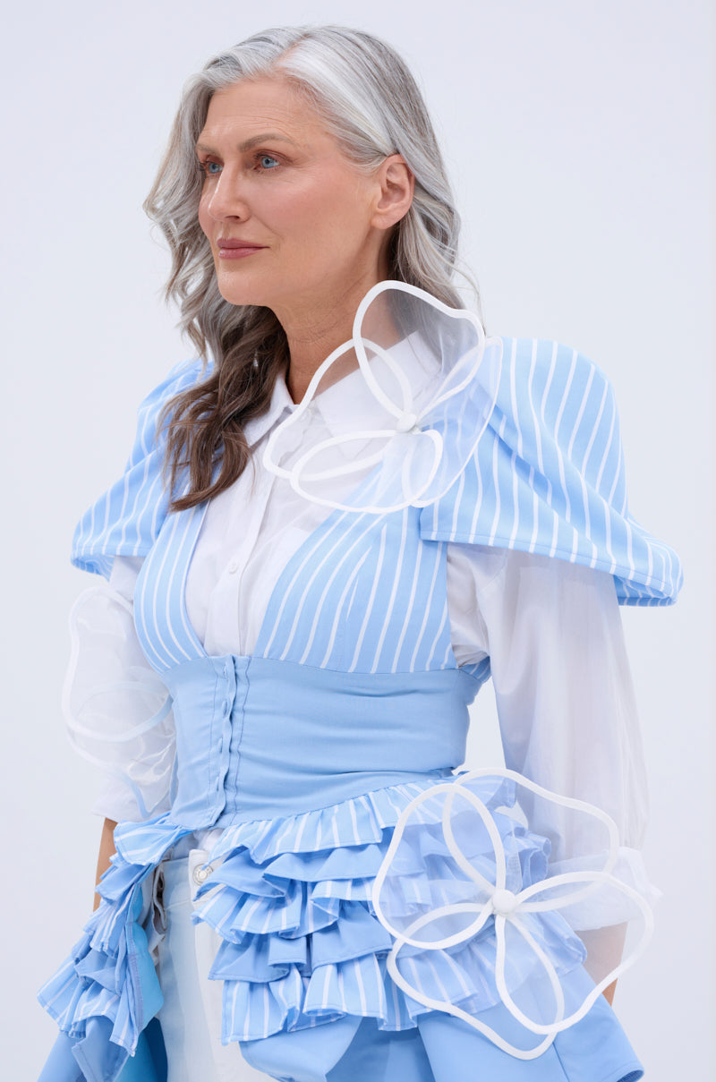 A woman with long gray hair wears a light blue and white striped dress with sheer mesh sleeves and large flower-shaped appliques, styled with the IN BLOOM BROOCH SET. She looks confidently to the side against a plain background. 0