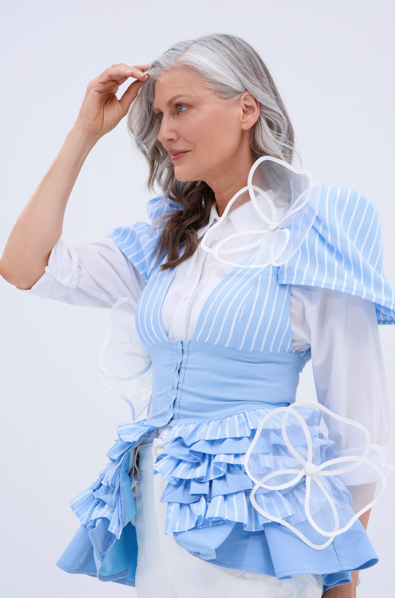 A woman with long gray hair stands against a plain light background, one hand near her head, wearing a stylish blue and white ruffled outfit adorned with the IN BLOOM BROOCH SET and sheer floral details for garden-inspired elegance. 1