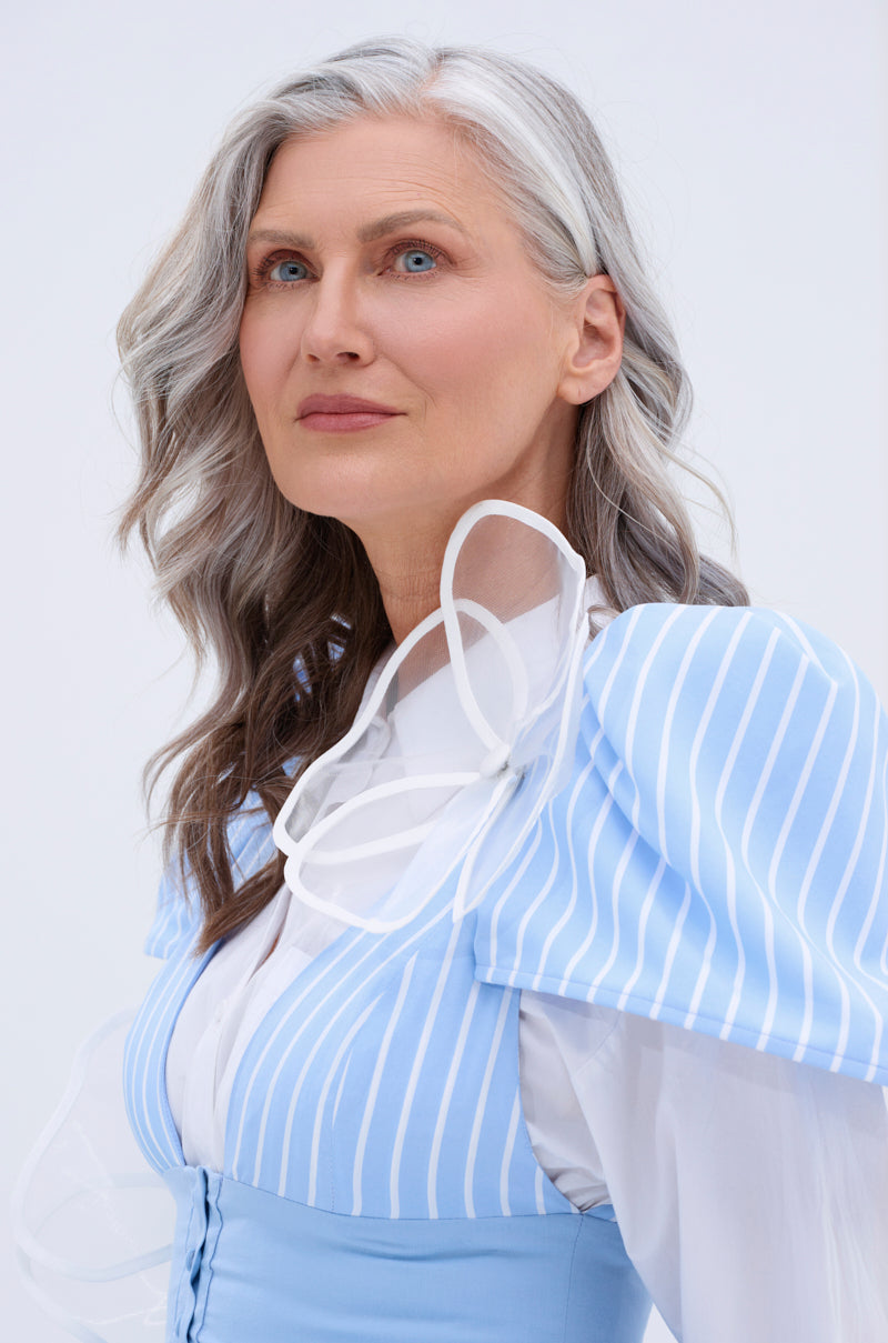 A woman with wavy gray hair and blue eyes wears the IN BLOOM BROOCH SET on a light blue and white striped dress, gazing confidently upward against a simple, light background. 2