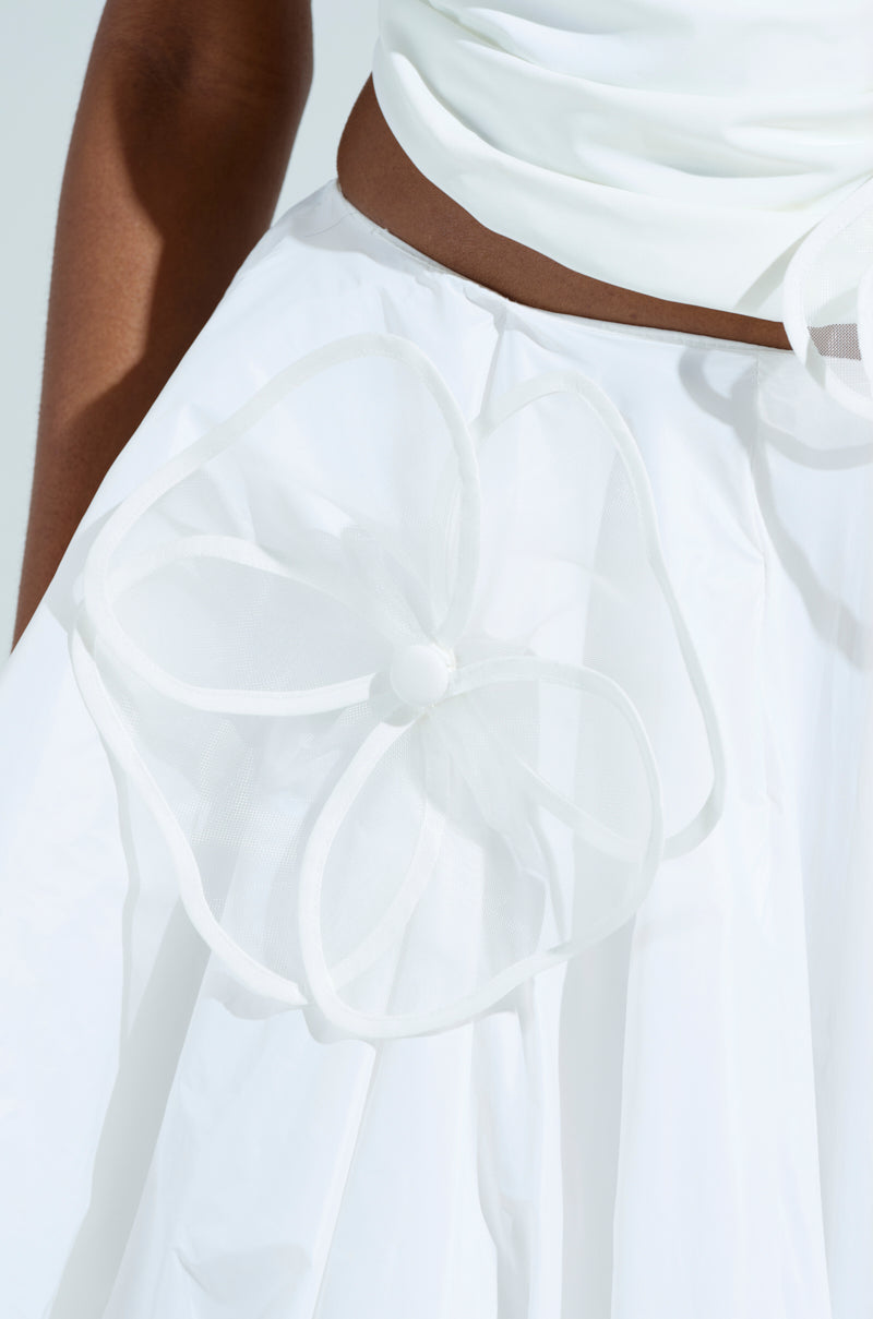 A woman in a white dress, highlighted by flower-shaped appliqués on the skirt of sheer unlined mesh, is featured close-up. The fabric's shiny texture enhances floral details, while a soft gradient background accentuates this elegant IN BLOOM BROOCH SET design. 6
