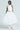 A person poses in an elegant, white one-shoulder dress with floral appliques on the shoulder and waist. The voluminous skirt pairs with unique white boots. The plain backdrop enhances the chic look of this ensemble, exuding a charming allure akin to the IN BLOOM BROOCH SET.