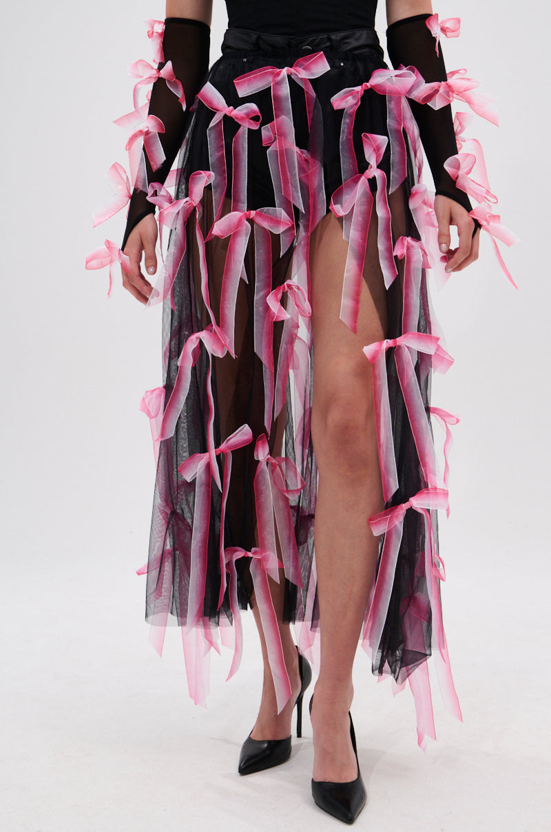 A person wears the BOW DOWN BOW SKIRT BELT—a sheer mesh black tulle skirt adorned with pink ribbon bows—paired with long black sleeves, also decorated with bows, and black high-heeled shoes. Skirt features a layered, partially see-through look.