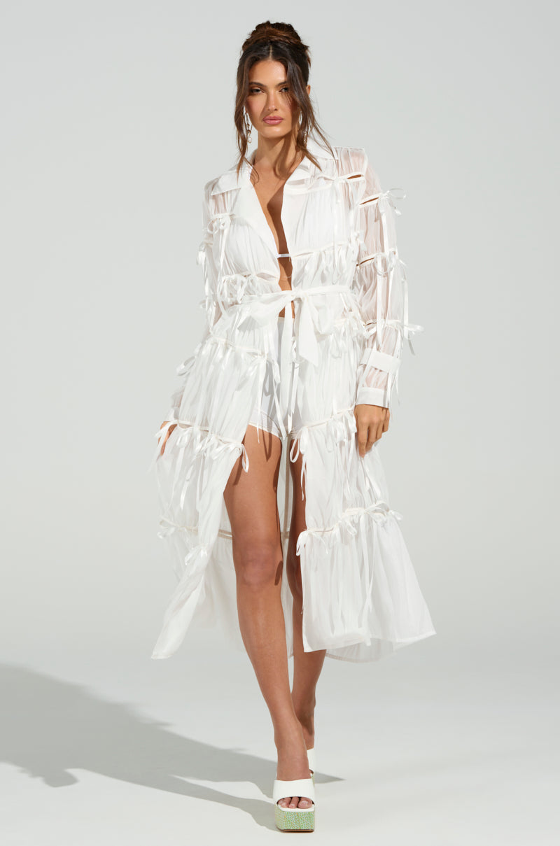 A woman poses in the RUBY CUTOUT ORGANZA TRENCH, a sheer white dress with bows, a deep neckline, and high slit. The outfit is accented by a self-tie belt and paired with white platform heels. Her hair is styled in a loose updo against a plain, well-lit background.