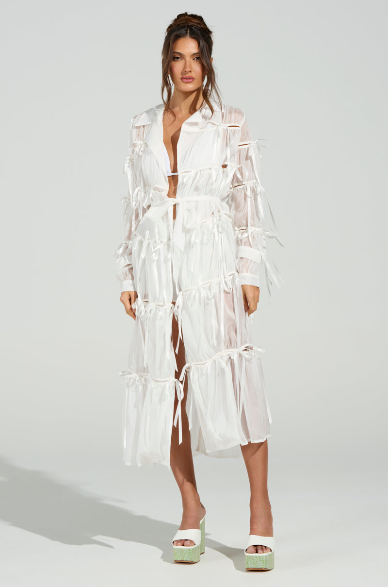 A woman in a chic RUBY CUTOUT ORGANZA TRENCH stands confidently against a neutral background. The trench boasts multiple bow ties and layers, enhanced with a self-tie belt, paired with platform heels. She gazes ahead, her hands resting by her sides.