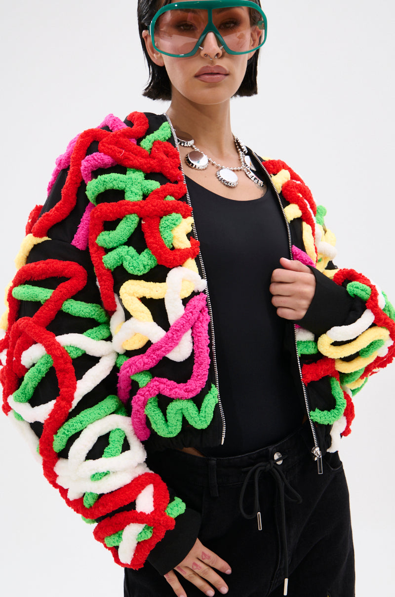 A person wears a black top and pants, chunky chain necklace, bold MOD 3D BOMBER jacket with a relaxed silhouette and thick multicolor appliqués in red, green, yellow, pink, white, plus large green-tinted glasses.