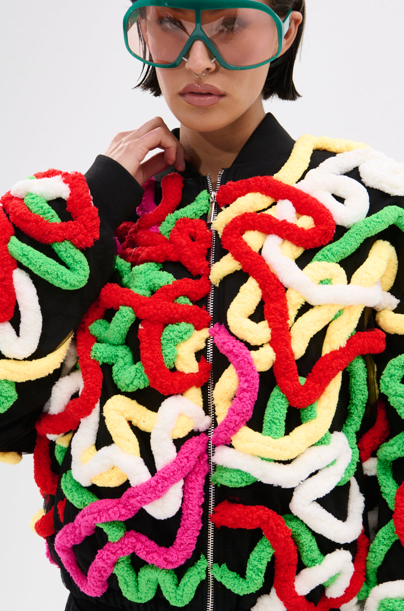 A person wears large green glasses and the MOD 3D BOMBER, a black jacket with thick, colorful yarn tufts in red, green, yellow, pink, and white. The multicolor appliqués form a bold, textured pattern.