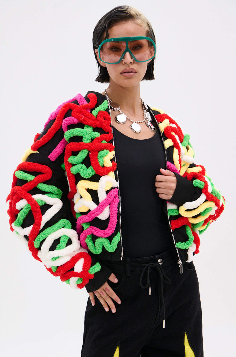 Wearing the MOD 3D BOMBER—a black top with colorful 3D appliqués and loops—paired with large green sunglasses and a chunky necklace, a person poses confidently against a plain background.