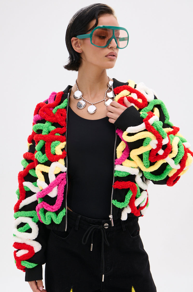 A woman in large teal sunglasses, a chunky silver necklace, and black top models the MOD 3D BOMBER, a relaxed fit jacket with red, green, white, yellow, and pink yarn-like loops against a plain light background.