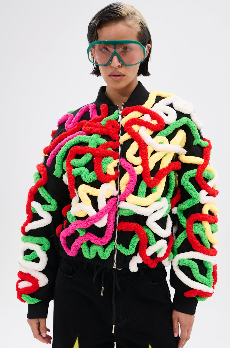 Wearing large green goggles and the MOD 3D BOMBER—relaxed fit, covered in thick, squiggly yarn appliqués in red, green, pink, white, and yellow—a person stands against a plain light background.