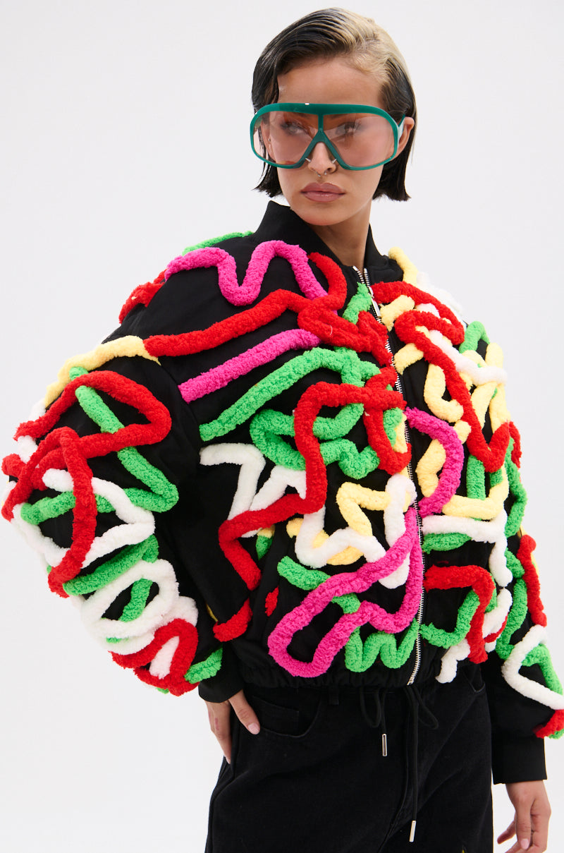 Wearing oversized green goggles and the MOD 3D BOMBER—featuring a relaxed fit, colorful chunky yarn squiggles, and multicolor appliqués—a person stands against a plain light background.