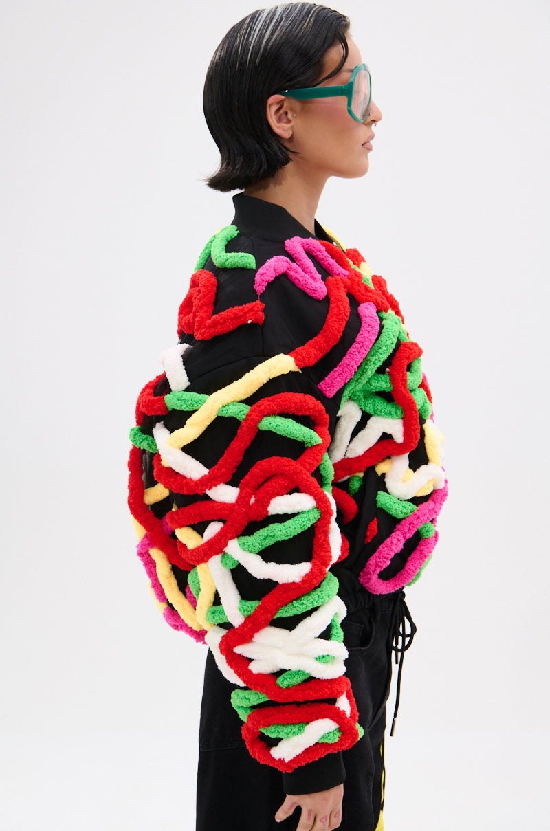 A person with short dark hair and large green glasses wears the MOD 3D BOMBER, featuring thick, colorful twisted yarn loops in red, green, white, yellow, and pink. The background is plain white.