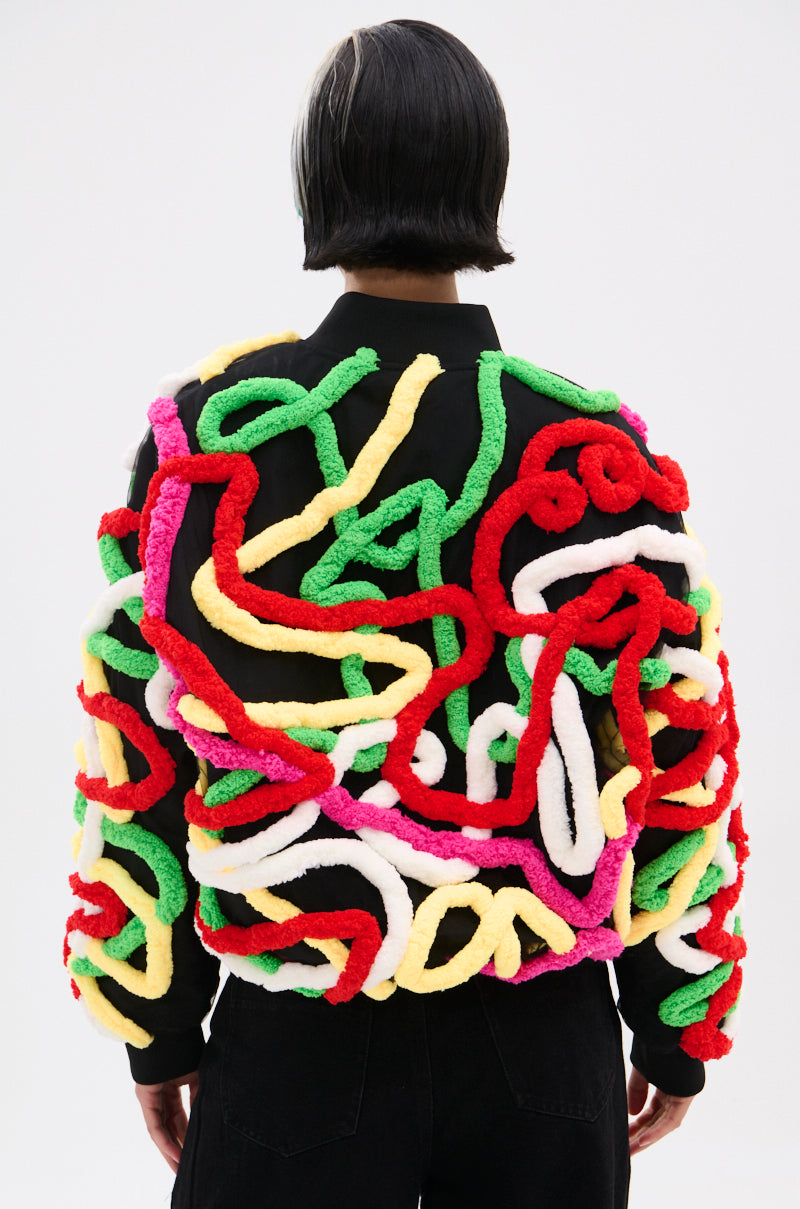 A person with chin-length black hair, seen from behind, wears the MOD 3D BOMBER—a relaxed-fit jacket adorned with thick, colorful squiggly yarn patterns in red, yellow, green, white, and pink. The background is plain and light.