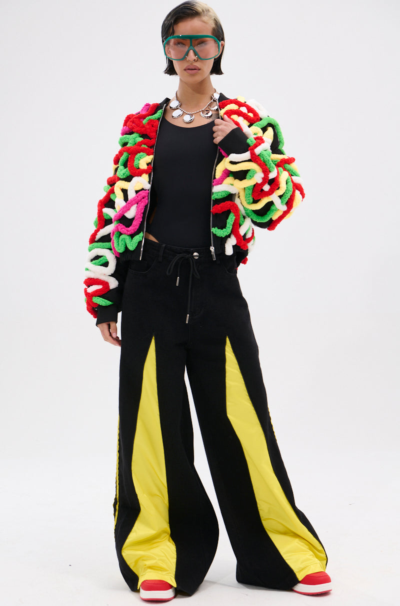 A person wearing the MOD 3D BOMBER—with multicolor appliqués and looping yarn—pairs it with a black top, wide-leg black pants with yellow panels, chunky sneakers, large sunglasses, and a silver necklace against a plain background.