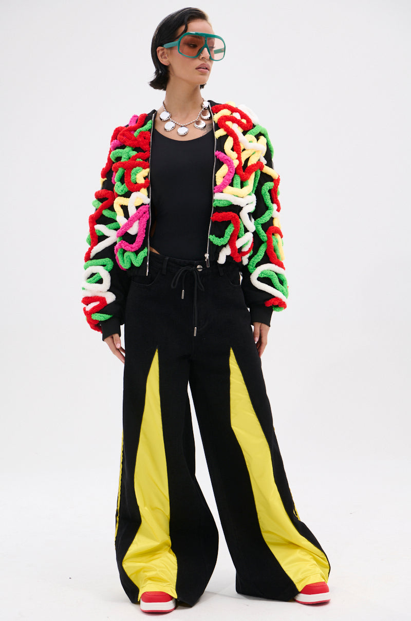 A person stands against a plain background wearing oversized green sunglasses, the MOD 3D BOMBER with multicolor appliqués, a black top, wide-leg black pants with yellow stripes, red-and-white shoes, and a silver statement necklace.