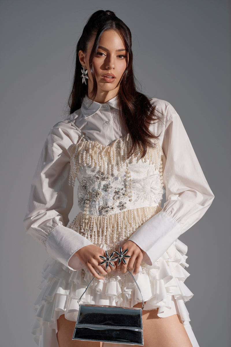 A woman poses in the BOLD FRINGE PEARL TOP, featuring ruffled details, pearlescent beads, and sequins over a collared shirt. She accessorizes with a shiny silver handbag, star-shaped jewelry, and soft waves in her long hair.