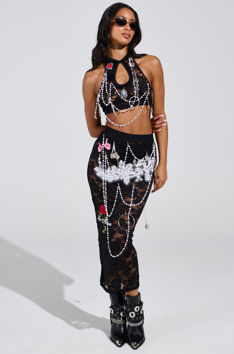 A woman poses confidently in a black lace dress with white embellishments and pearl chains, wearing black sunglasses, a halter neck top, and the CONTROLLED CHAOS LACE MIDI SKIRT. Her fitted long skirt has a high-rise silhouette, complemented by black boots with silver buckles.
