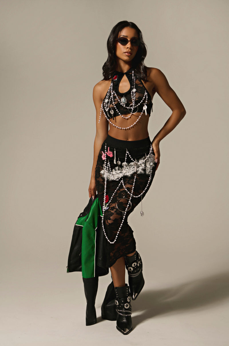 Dressed in a CONTROLLED CHAOS LACE MIDI SKIRT, featuring sheer black fabric adorned with colorful patches and pearls, a person with long dark hair, sunglasses, and black boots confidently holds a green and black jacket. The plain backdrop enhances their striking style.