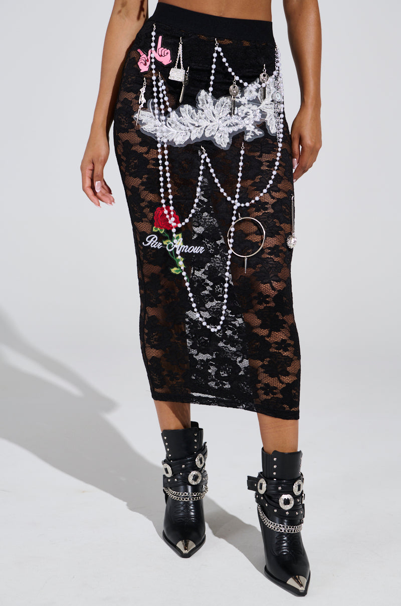 A person is wearing the CONTROLLED CHAOS LACE MIDI SKIRT, a stretchy black midi skirt with pearls, lace patches, silver accents, a chain belt, and "amour" near a red rose detail. They're also in black studded boots.