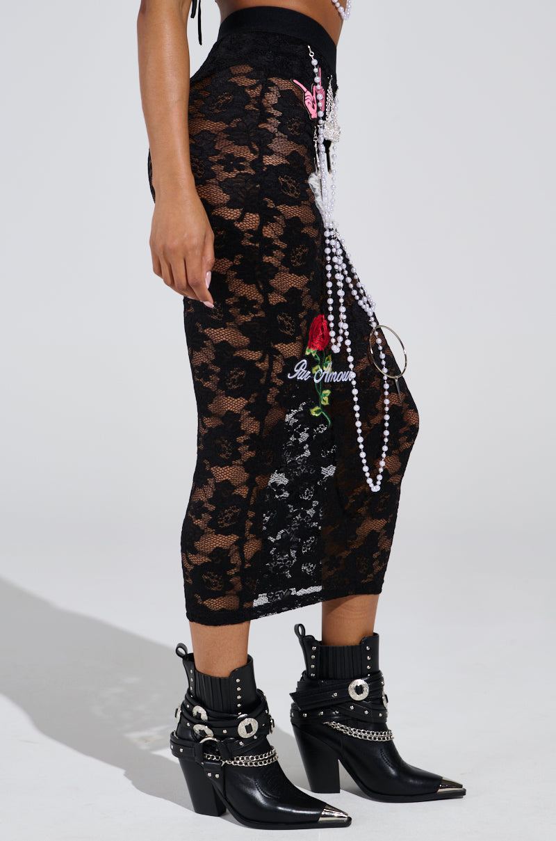 Wearing the CONTROLLED CHAOS LACE MIDI SKIRT, featuring black lace with pearls and floral embroidery, paired with black leather ankle boots adorned with silver buckles and studs, a person stands against a plain, light-colored backdrop.