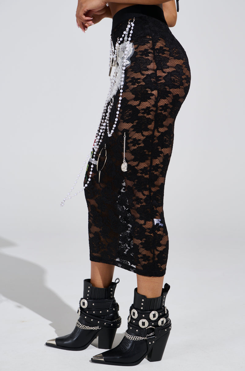 Sporting the high-rise black CONTROLLED CHAOS LACE MIDI SKIRT adorned with chains and pearls, they pair it with pointed black boots featuring silver studs, buckles, and decorative elements against a plain white background.