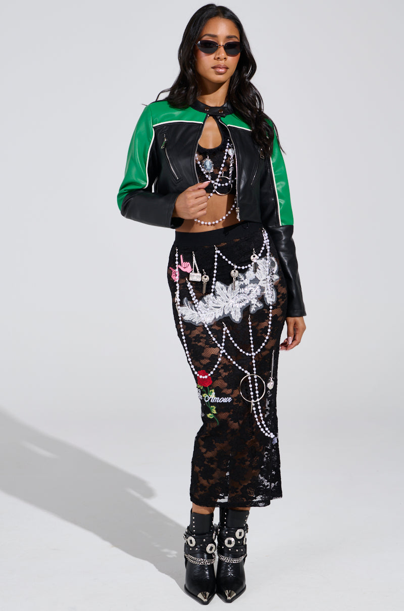 A woman confidently stands against a plain background, wearing sunglasses, a black and green leather jacket, the CONTROLLED CHAOS LACE MIDI SKIRT with intricate white patterns, and black boots with silver embellishments that complete her high-rise silhouette.