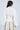A woman with long brown hair, shown from behind, wears the VERA WOVEN TRIM BLOUSE with balloon sleeves and a back zipper, paired with pearl earrings and a light-colored textured skirt.