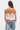 A woman with long brown hair and hoop earrings wears the GOING PLACES CORSET WITH TWEED, a pink lace-up top with multicolored trim and subtle peplum detail, paired with blue jeans with white floral embroidery and a small shoulder tattoo.