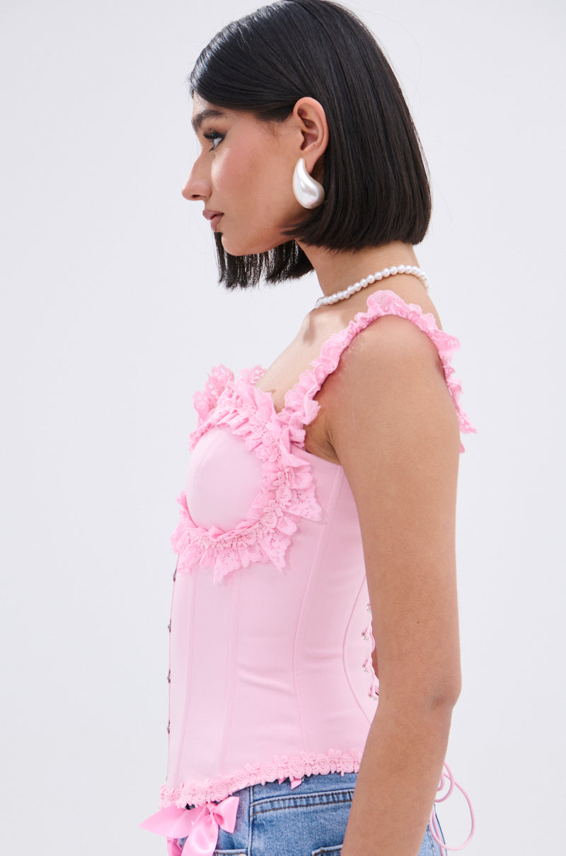 A woman with a bob haircut wears the STUCK IN A DREAM CORSET TOP in light pink with lace trim, blue jeans, pearl necklace, and large white earrings, posing in profile against a plain light background.