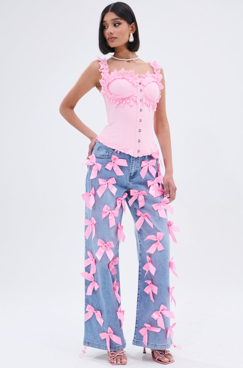 A woman models the STUCK IN A DREAM CORSET TOP, a fitted pink piece with lace trim, paired with blue jeans featuring large pink bows. She finishes the look with pink heels and a pearl necklace against a plain white background.