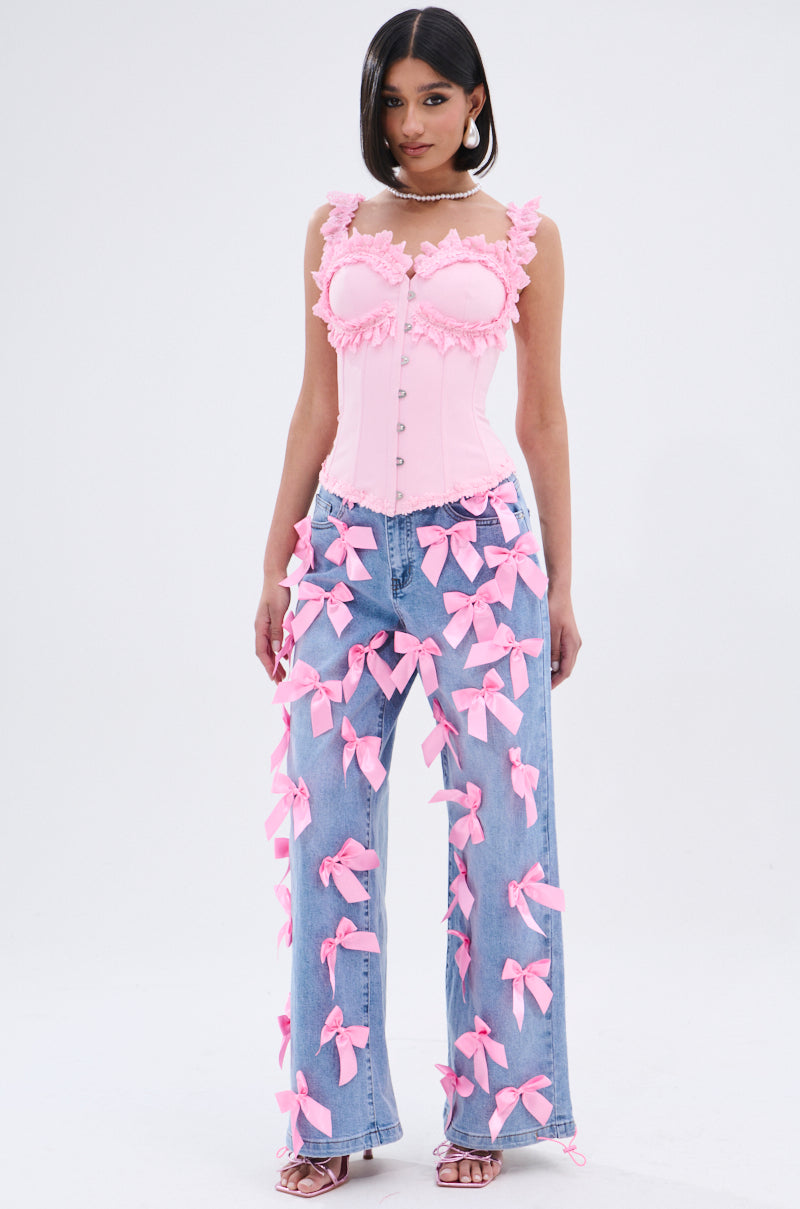 A woman stands against a white background wearing the STUCK IN A DREAM CORSET TOP, a light pink corset with a sweetheart neckline and delicate lace trim, paired with light blue wide-leg jeans accented with pink bows. She has straight hair and wears sandals.
