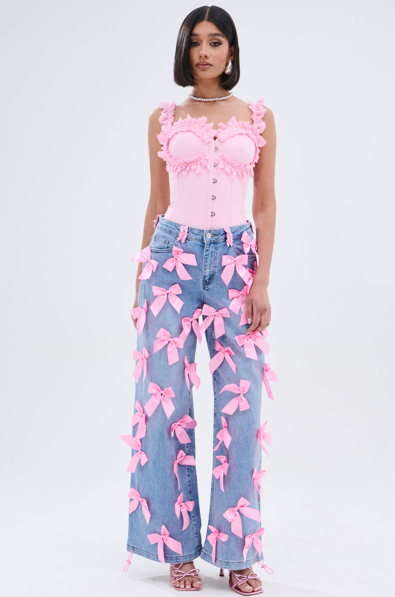 A woman wears the STUCK IN A DREAM CORSET TOP, a pink piece with a sweetheart neckline, pearl accents, and lace trim. She pairs it with high-waisted blue jeans decorated with large pink bows, straight dark hair, and pink heels.