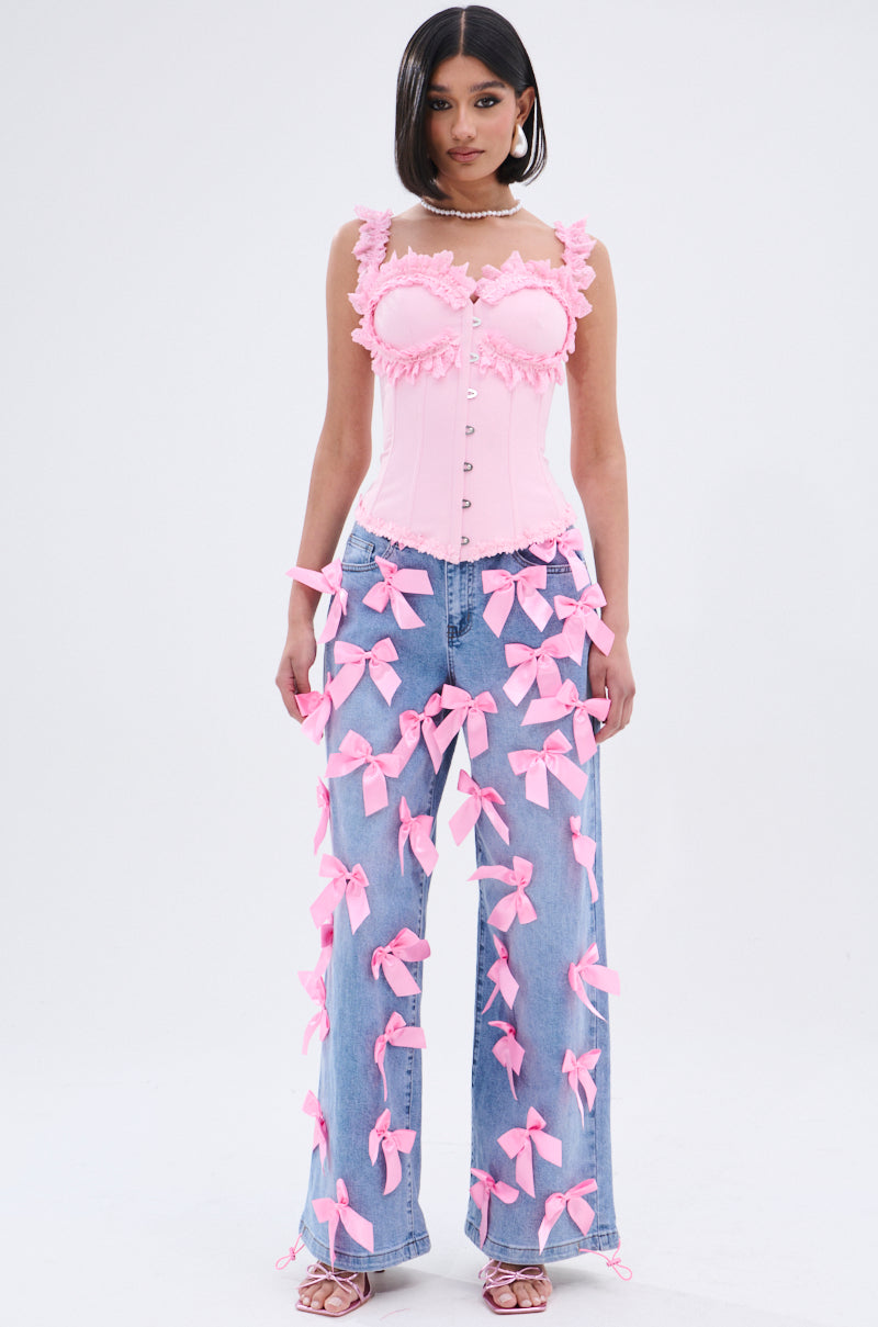 A woman faces forward wearing the STUCK IN A DREAM CORSET TOP in light pink with ruffles, a sweetheart neckline, and lace trim, styled with loose blue jeans adorned with pink bows, strappy pink heels, and straight shoulder-length hair.