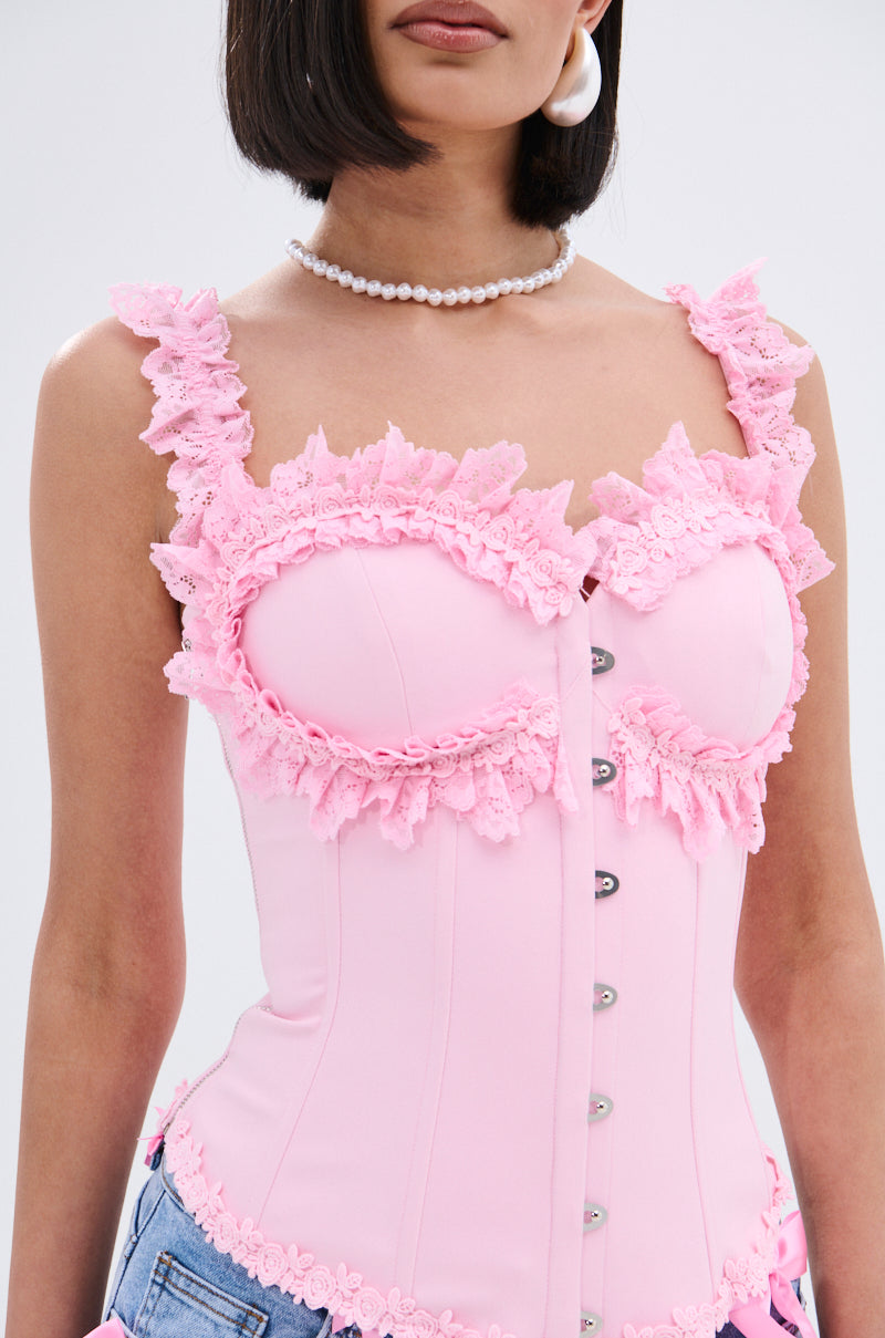 A woman wears the STUCK IN A DREAM CORSET TOP, a light pink piece with lace trim and pearl buttons, styled with blue jeans, a pearl necklace, large white earrings, and straight shoulder-length hair.