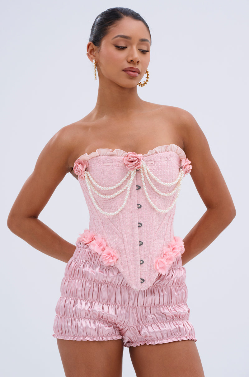 A woman wears the SOMEDAY WELL KNOW CORSET TOP in pale pink, featuring a sweetheart neckline, floral and pearl embellishments, and ruffles, paired with matching high-waisted ruched shorts. Her hair is tied back and gold hoop earrings shine against a neutral background.