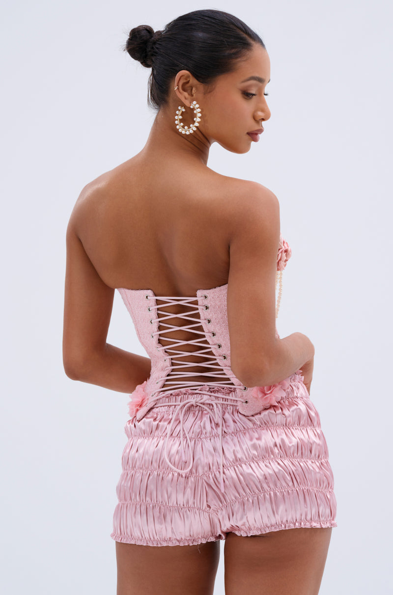A woman with a neat bun wears pearl hoop earrings and the SOMEDAY WELL KNOW CORSET TOP, featuring a pink sweetheart neckline and lace-up back, paired with shiny ruched pink shorts. She poses with her back and side to the camera.