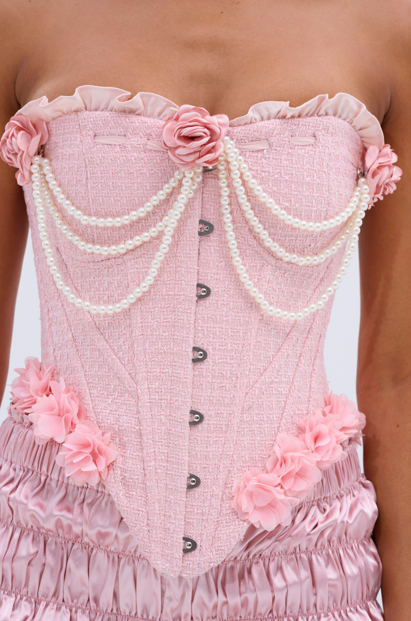 A person wears the SOMEDAY WELL KNOW CORSET TOP, a pink corset with a sweetheart neckline, fabric flowers, white pearl draping over the chest, paired with matching pink ruffled shorts for an elegant and feminine look.