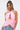 A woman with long dark hair in a high ponytail models the FEATHER QUEEN BLOUSE, a sleeveless pink feather halter top with keyhole neckline and open back, styled with light blue jeans and statement earrings against a plain backdrop.